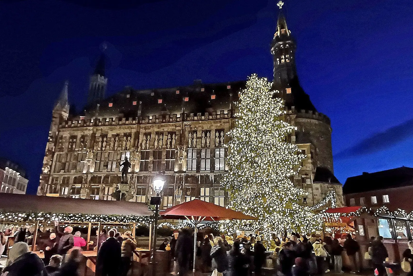 Het verlichte stadhuis van Aken met een grote kerstboom en de kramen van de kerstmarkt bij avond